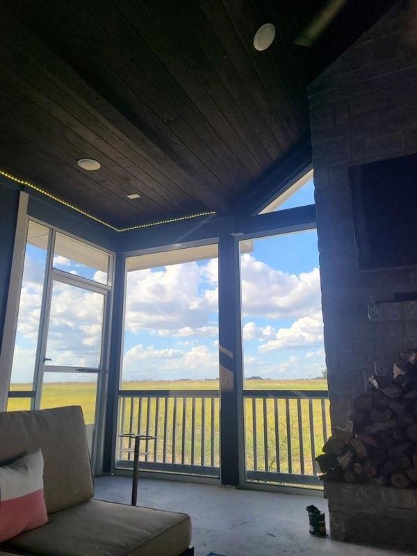A sunlit screened-in porch with wood ceiling, stone fireplace with firewood, and a view of a grassy field under a blue sky.
