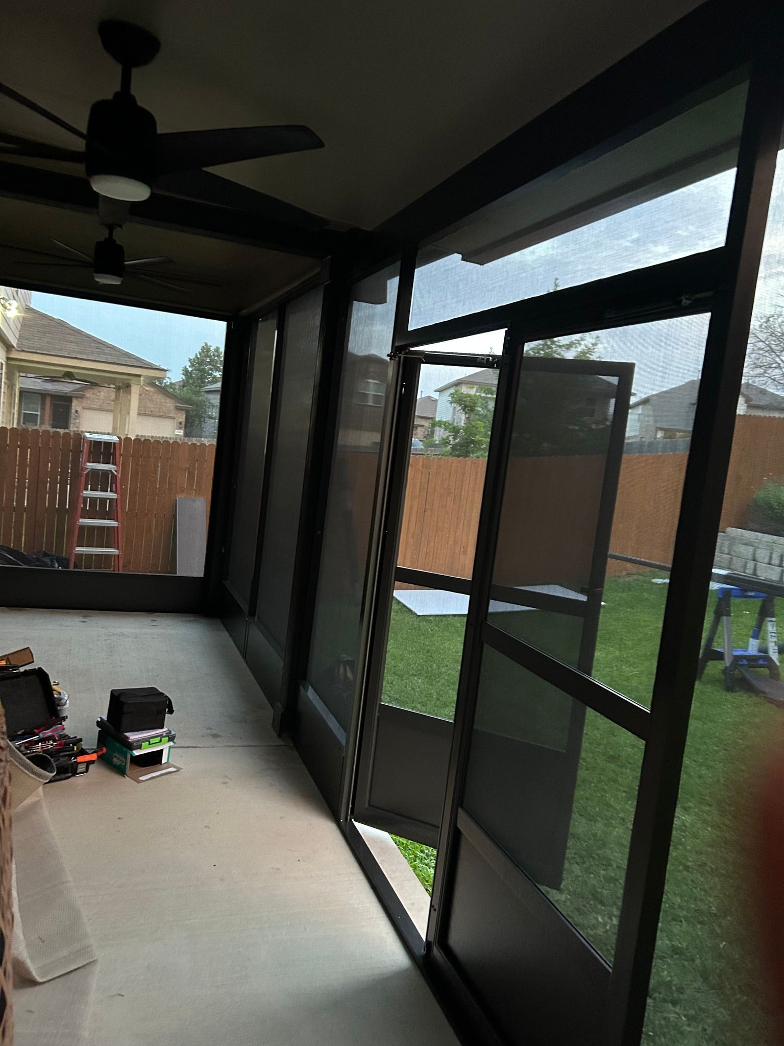 An interior view of a screened-in porch with dark frames, ceiling fans, and a view of a backyard with a wooden fence.