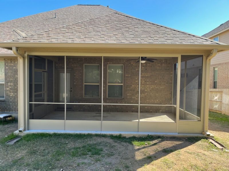 A screened-in patio attached to a tan brick house with a brown shingled roof under a clear blue sky.