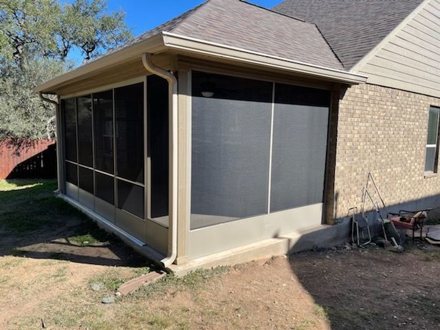 A beige screened-in porch addition attached to the side of a brick house with a brown shingled roof.
