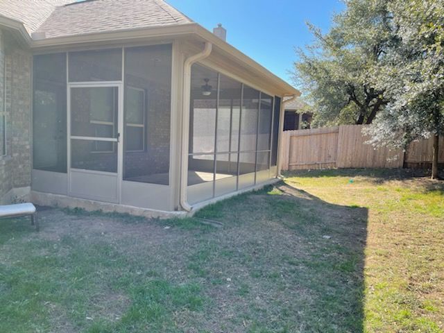 A tan screened-in porch attached to a brick house, overlooking a grassy backyard with a wooden fence and trees.