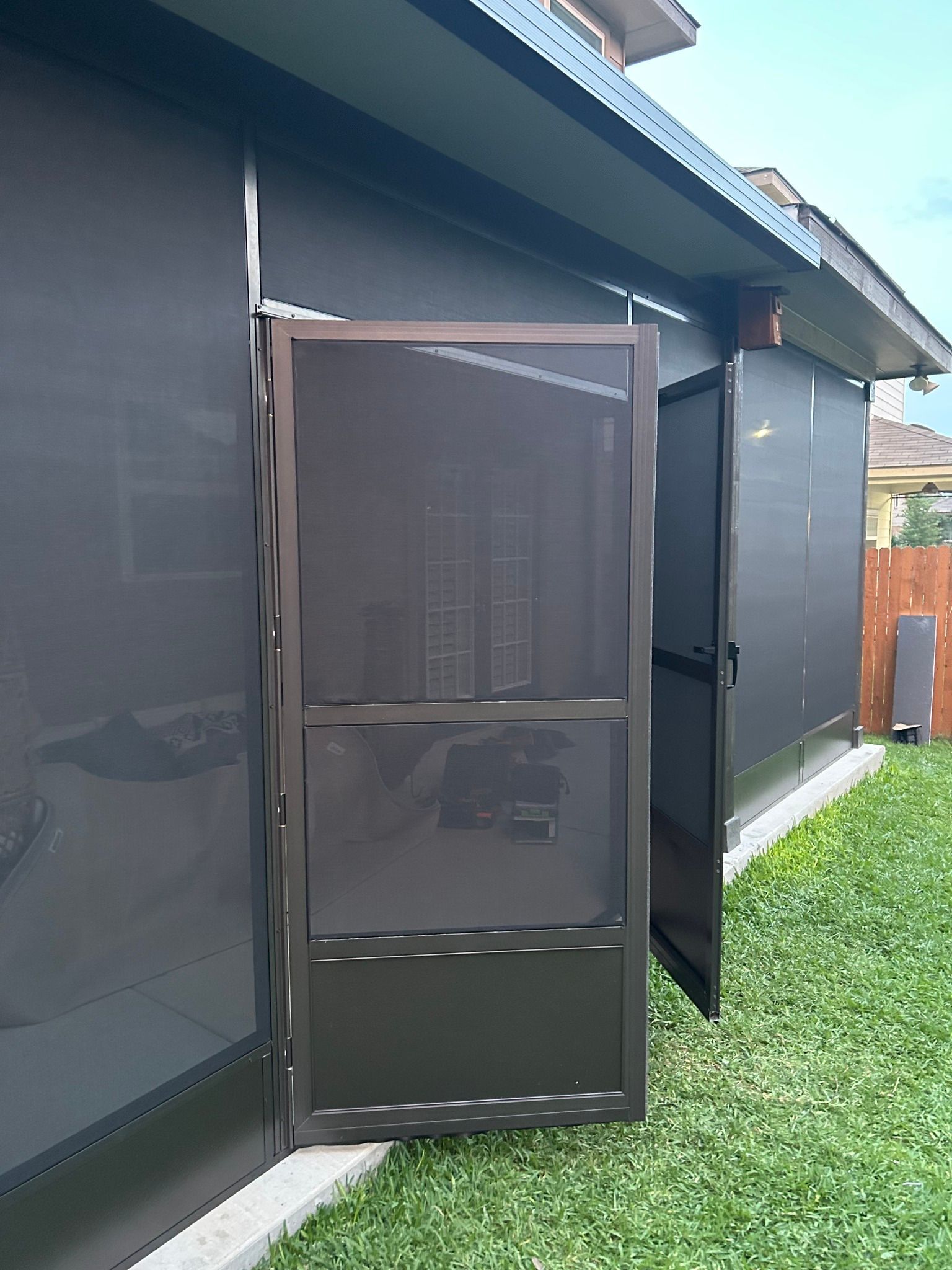 A brown framed screen door stands open, revealing a patio enclosure with mesh screening, set against a green lawn.