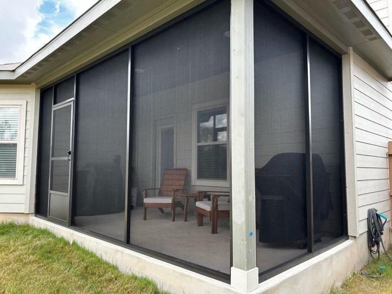 A screened-in porch with a metal door, outdoor seating, and a grill, attached to the side of a house.