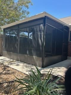 A screened-in patio enclosure with dark mesh panels and a white roof sits on a concrete slab in a sunny yard.
