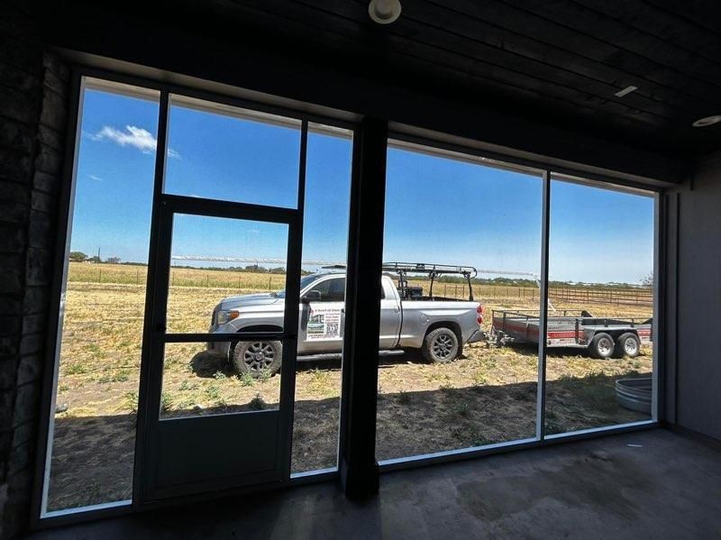 A view through floor-to-ceiling windows showing a pickup truck with a ladder rack and a utility trailer on a grassy lot.