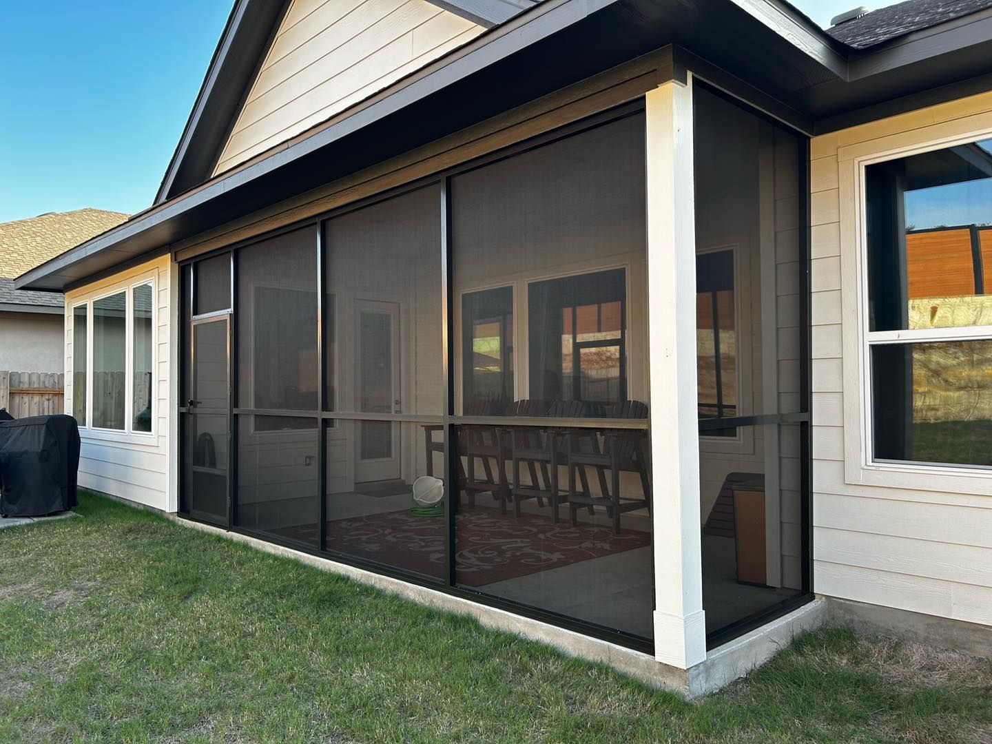 A backyard patio enclosed with black screen walls, attached to a house with cream-colored siding and a dark trim roof.