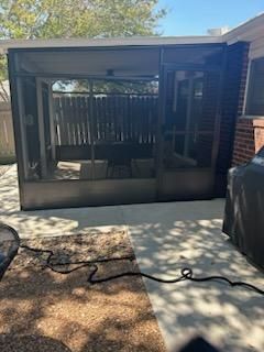 A dark-framed screened-in porch with outdoor furniture stands on a concrete patio next to a brick house exterior.