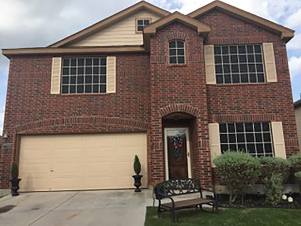 A two-story red brick house with a tan garage, beige shutters, a front door, and a small bench on a green lawn.