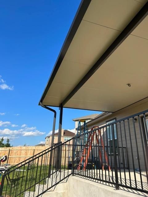 Low-angle view of a patio cover with a black railing, a downspout, and an orange ladder against a house under a blue sky.