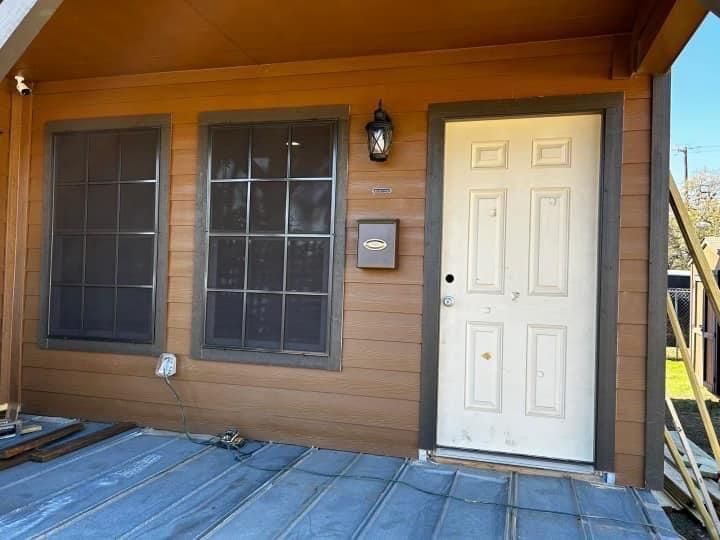 A beige door and two brown-trimmed windows on a wood-sided house, viewed from a porch under construction.