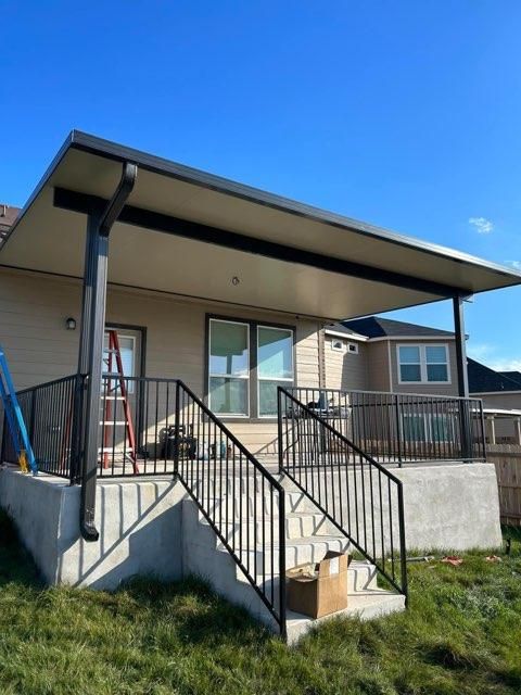 A covered concrete patio with black metal railings and stairs, attached to the back of a tan house under a blue sky.