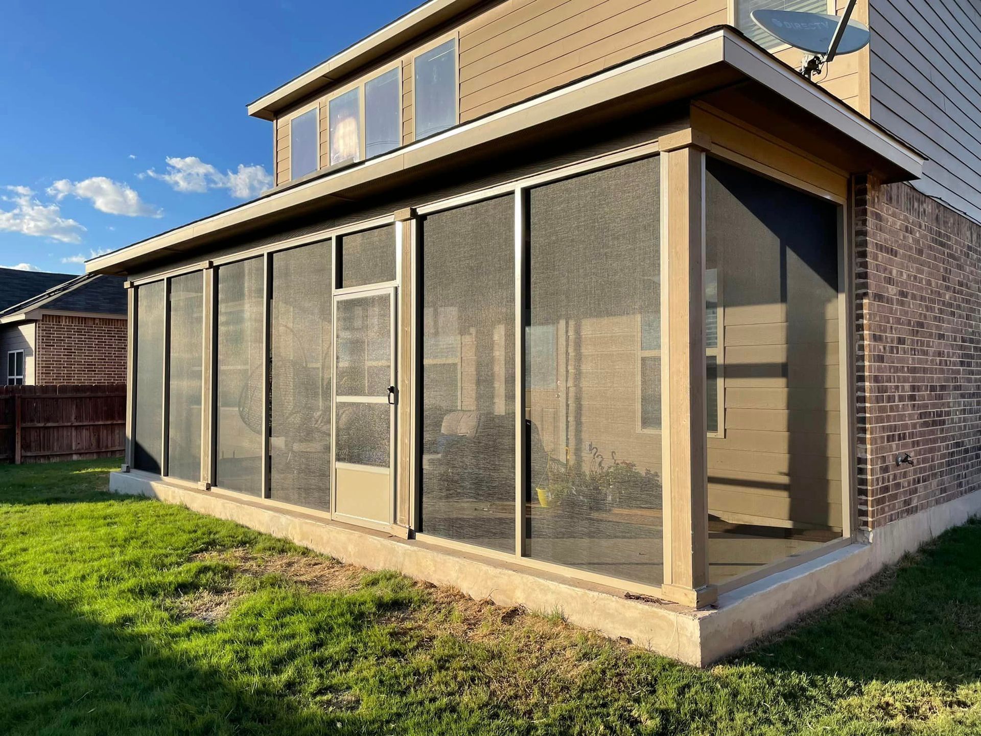 A beige screened-in porch attached to the back of a brick two-story house with a green lawn under a sunny blue sky.
