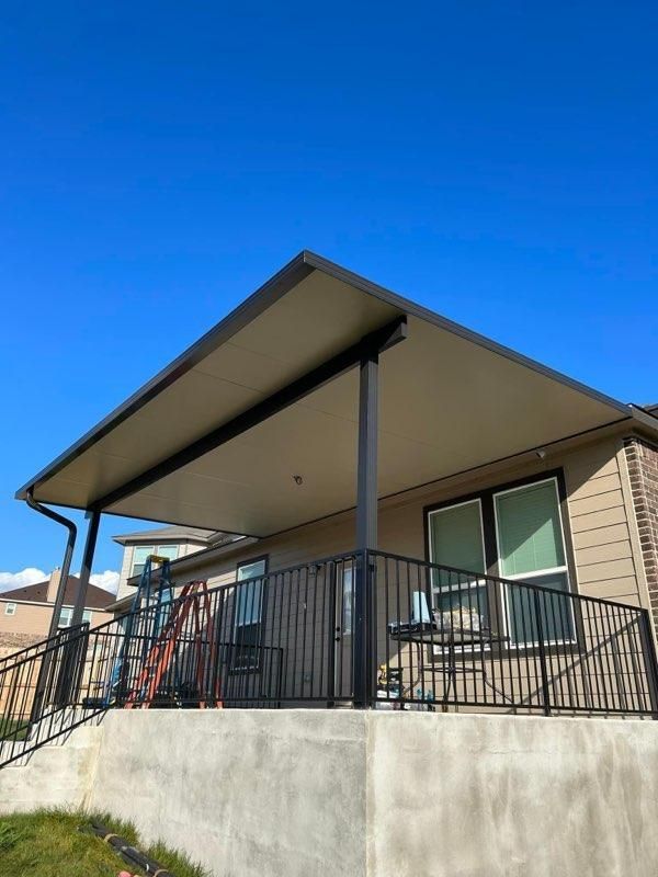 A tan house with a dark metal patio cover, black iron railings, and a concrete porch under a clear blue sky.