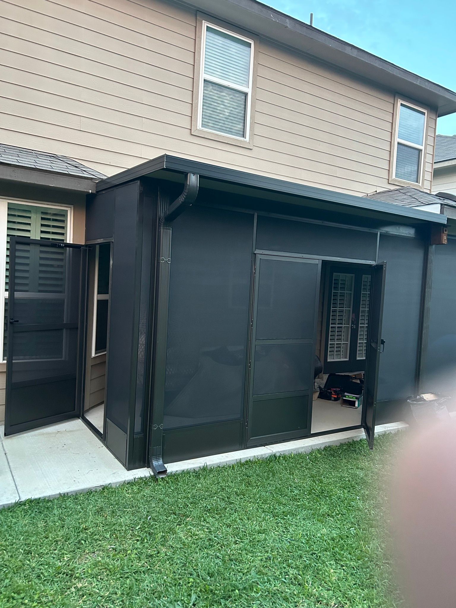 Exterior view of a house with a dark screen enclosure over the patio, featuring a partial open door and a grassy lawn.