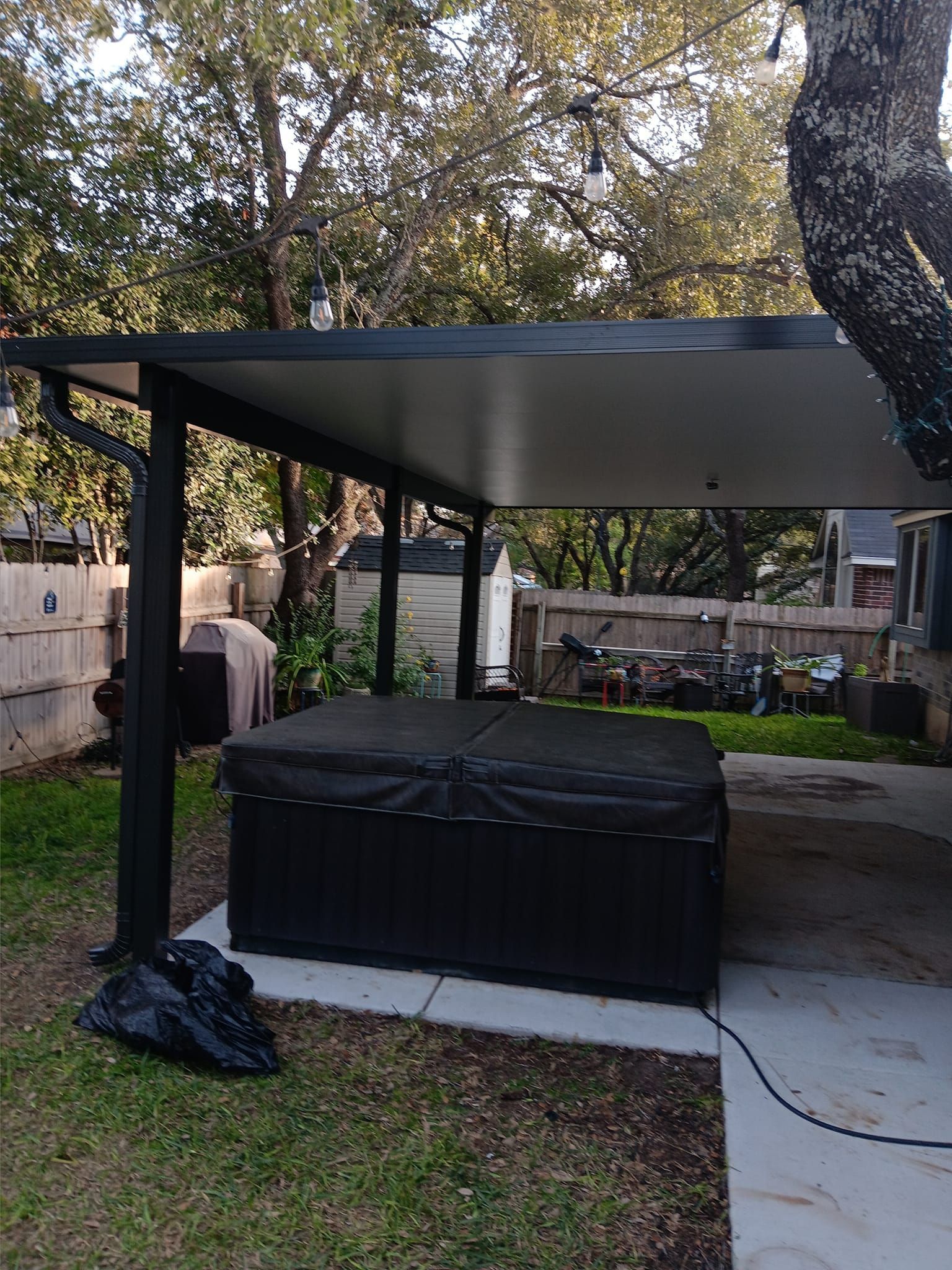 A black hot tub with a cover sits under a modern, flat-roofed metal patio cover in a backyard setting.