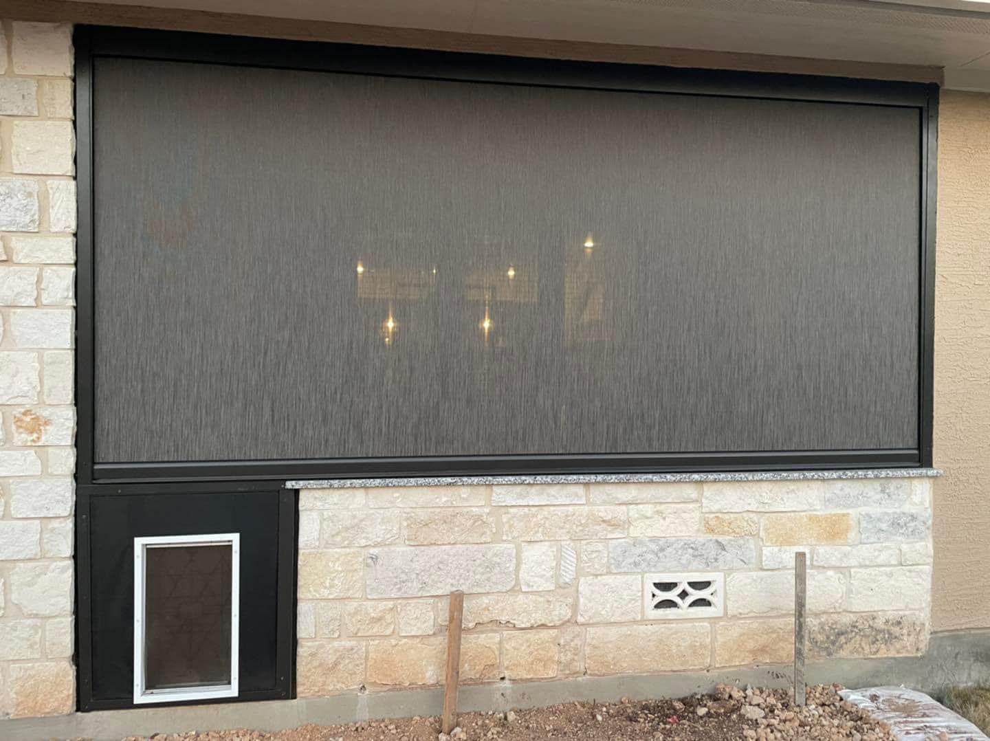 A black pet door installed in a stone wall beneath a large outdoor sun shade screen.