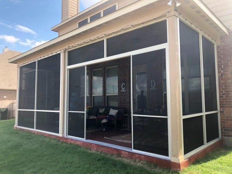 A beige, screened-in patio enclosure with white framing attached to a brick house, overlooking a green lawn.