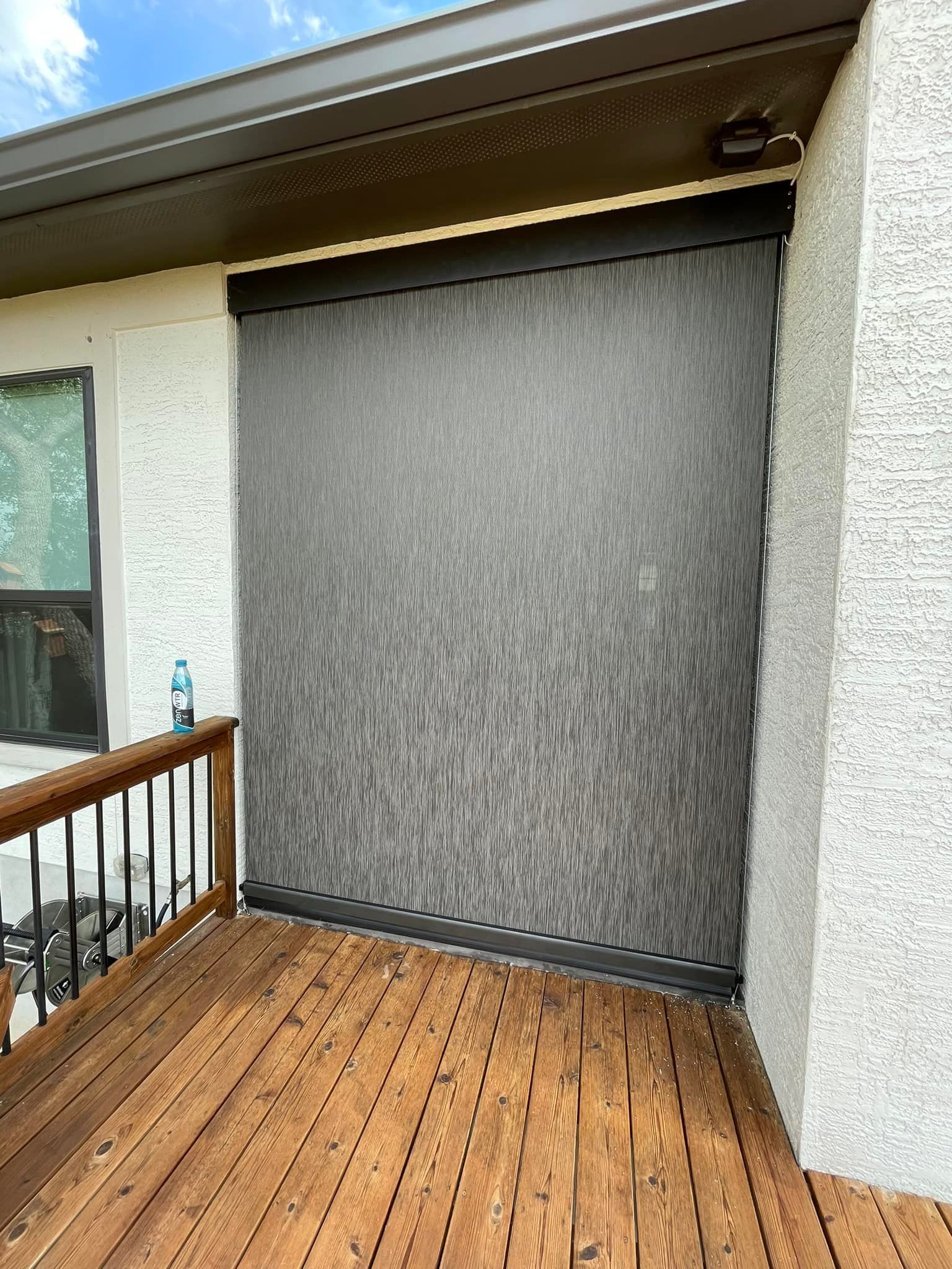 A dark grey outdoor solar shade mounted on a patio wall above a wooden deck with a railing.