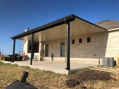 A beige stone house with a modern, flat-roofed black patio cover over a concrete slab on a sunny day.