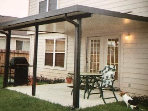 A covered patio area attached to a house with outdoor furniture, a grill, and a dog on the grass.