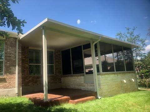 A covered patio with a screened-in enclosure attached to a brick house on a sunny day with green grass in the foreground.