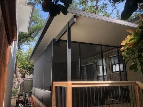 A screened-in patio with a dark metal frame, light-colored roof, and wooden railing, viewed from an outdoor angle.