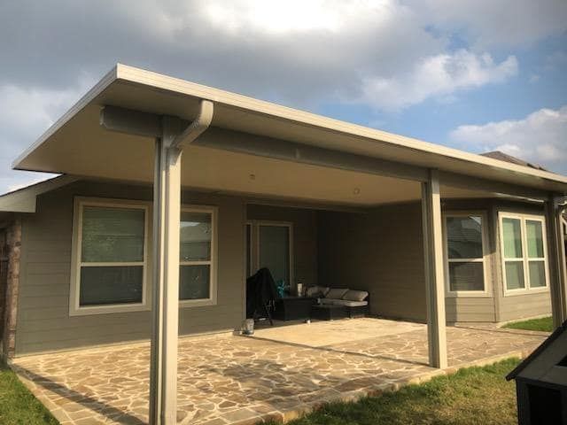 A covered stone patio attached to a gray house, featuring a large roof overhang supported by two tan vertical posts.
