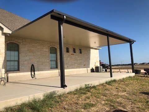 A tan stone house features a large, covered concrete patio with a dark roof and black support posts on a sunny day.