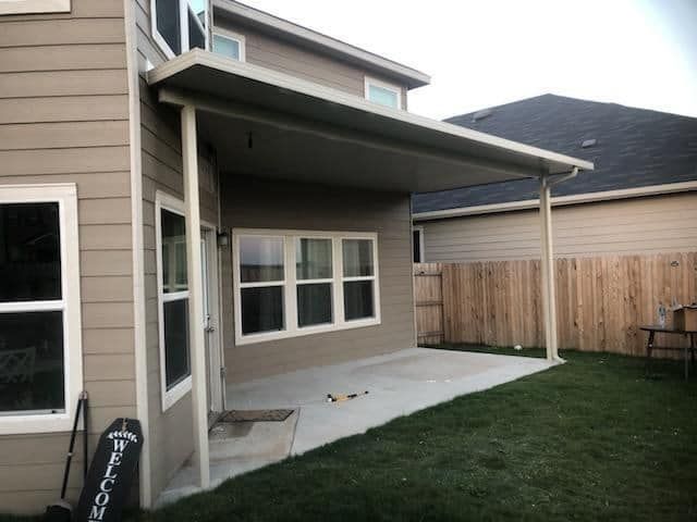 A back patio of a beige house with a covered concrete slab, three-pane windows, and a grassy yard with a wood fence.