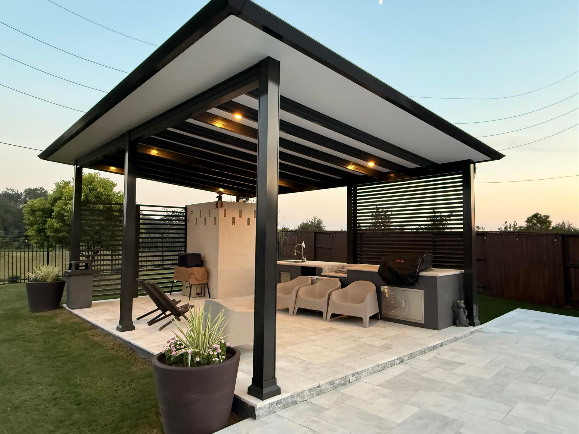 A modern, covered outdoor kitchen and lounge area featuring a stone structure, seating, and decorative black privacy panels.