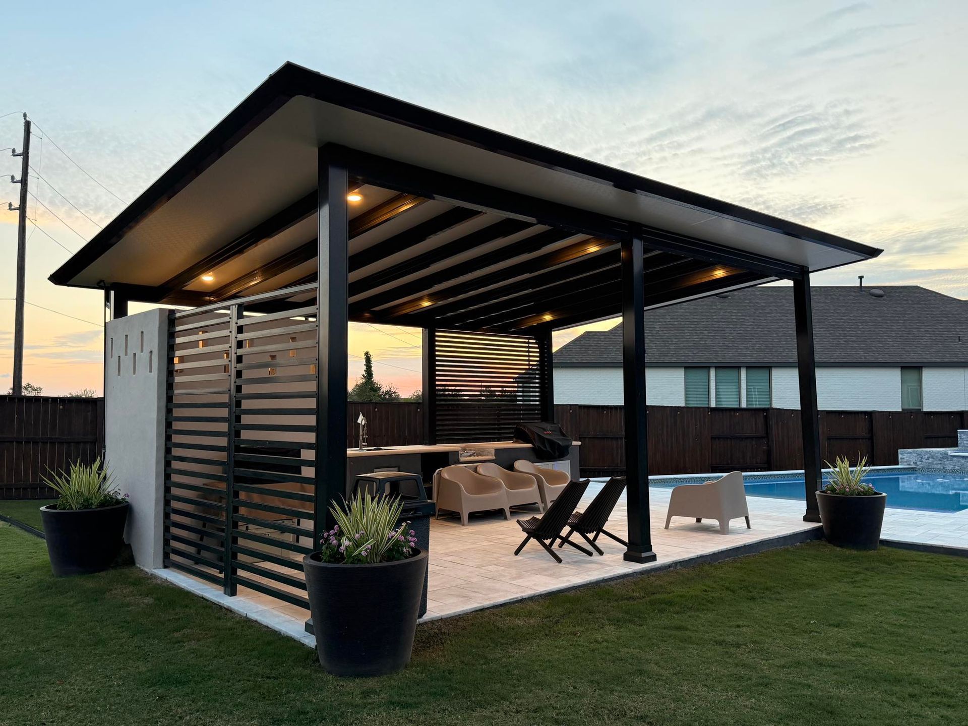 A modern black pergola patio with an outdoor kitchen, furniture, and pool in a backyard at sunset.