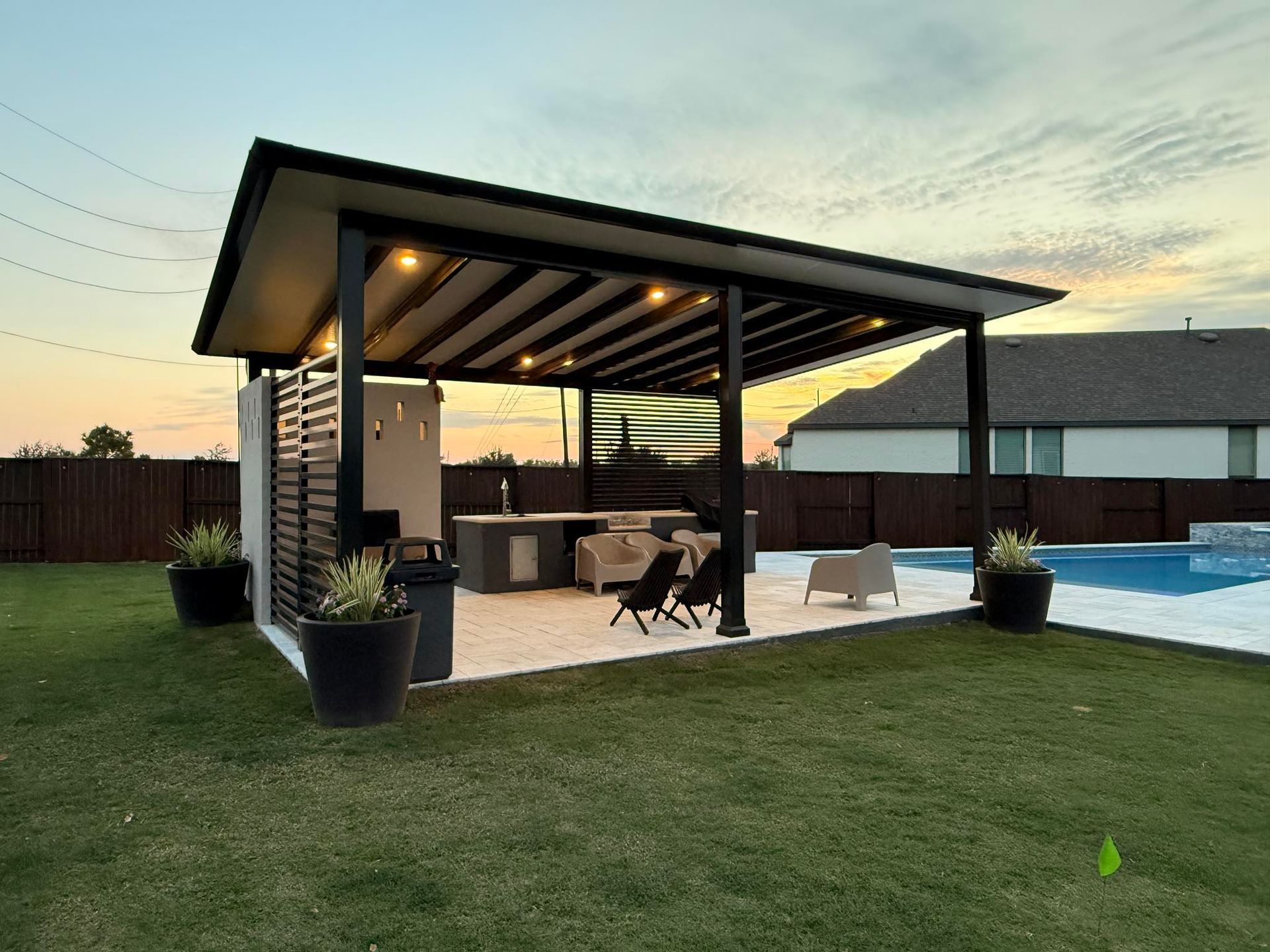 A modern covered outdoor patio with a kitchen, lounge seating, and recessed lighting, situated next to a swimming pool.