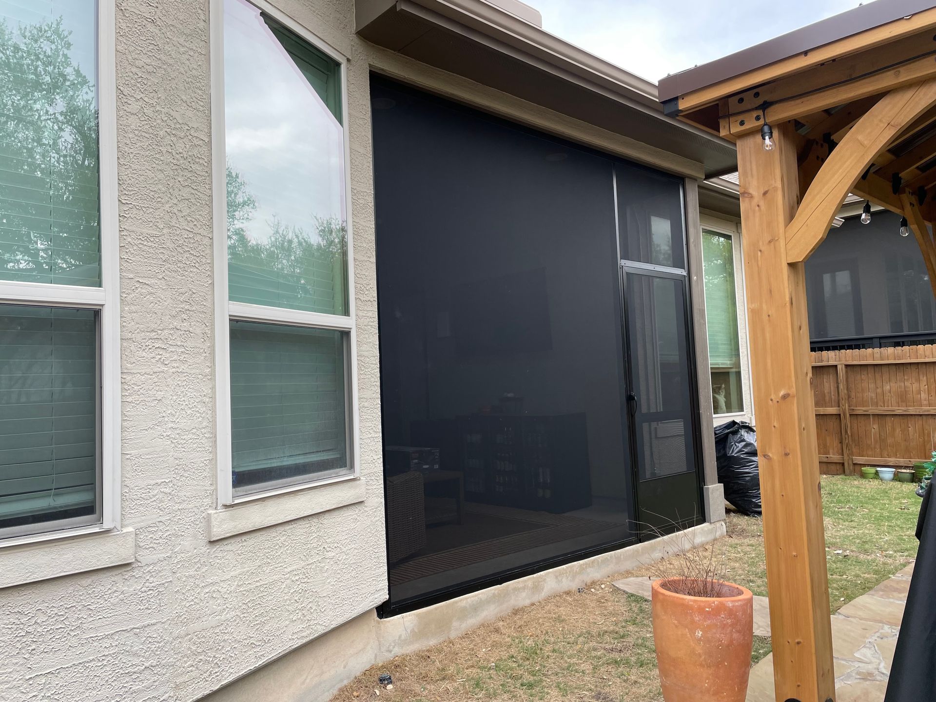 A black mesh patio privacy screen installed on a residential house exterior near a wooden gazebo structure.