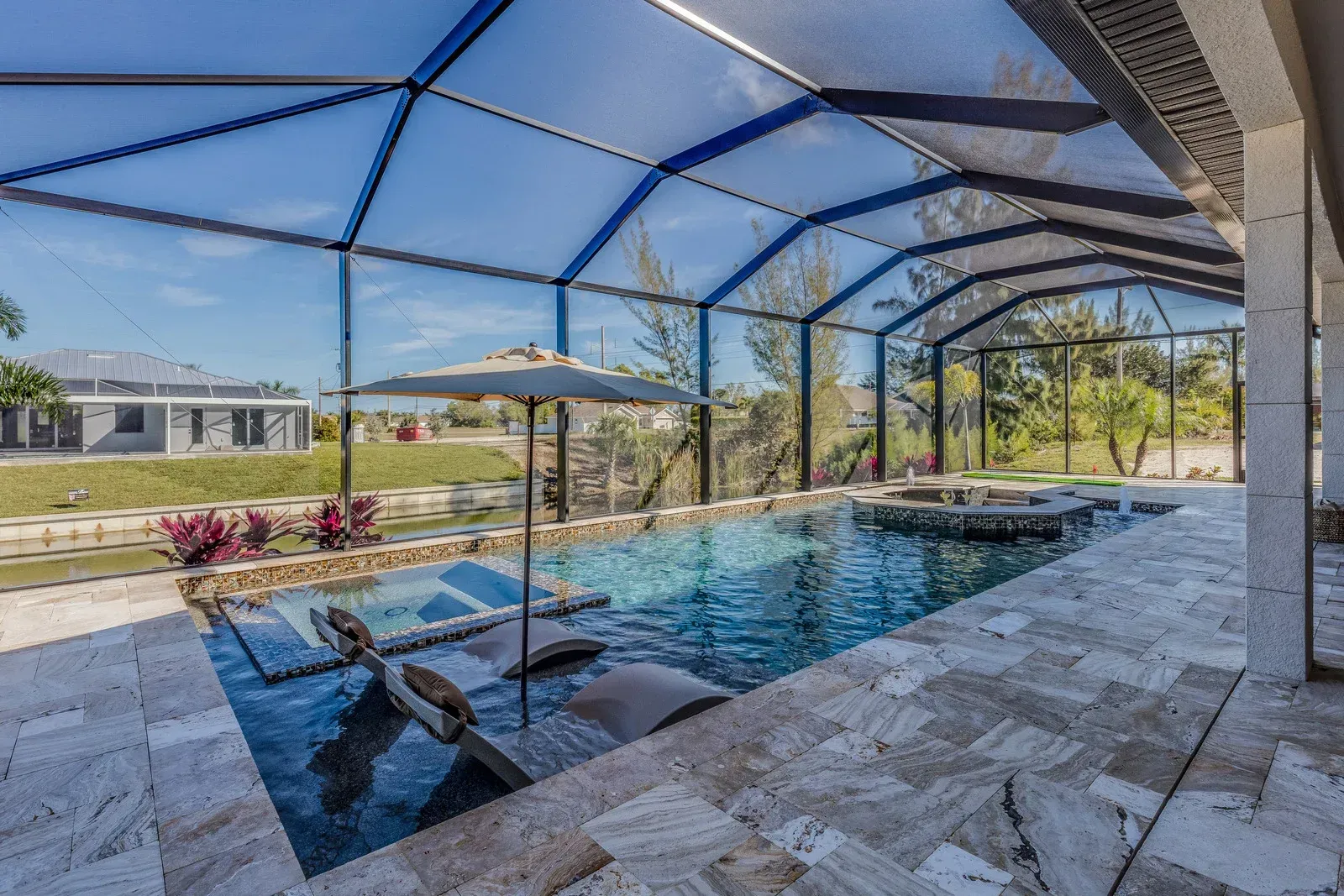 A screened-in patio features a swimming pool with submerged lounge chairs, a built-in spa, and light-colored stone pavers.