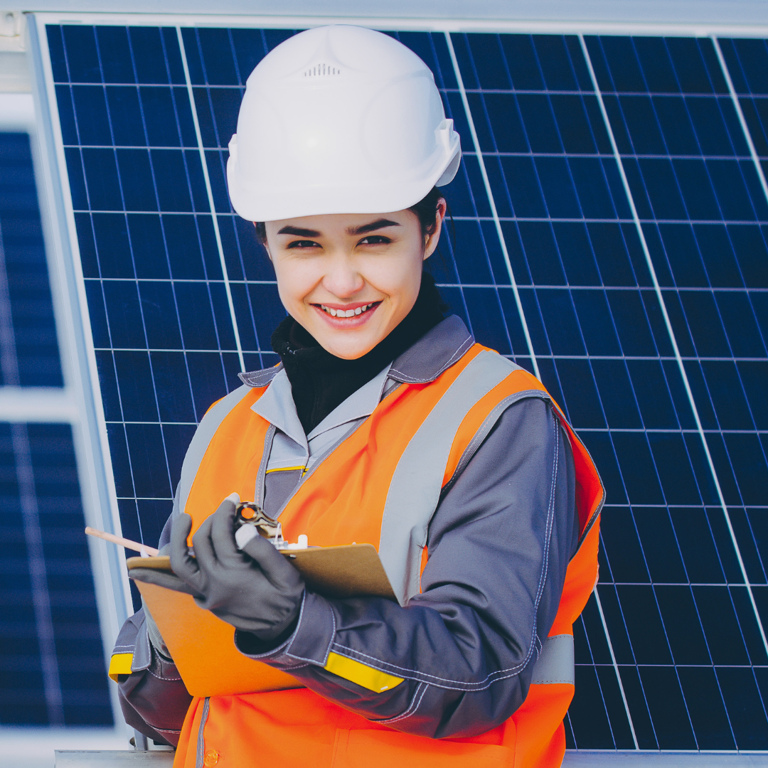 A woman wearing a hard hat is holding a clipboard in front of a solar panel.