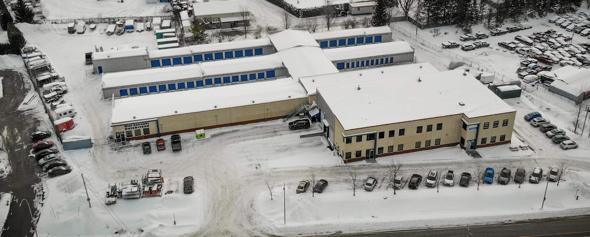 Vue aérienne de bâtiments industriels enneigés, avec leurs parkings, leurs véhicules et les arbres environnants.