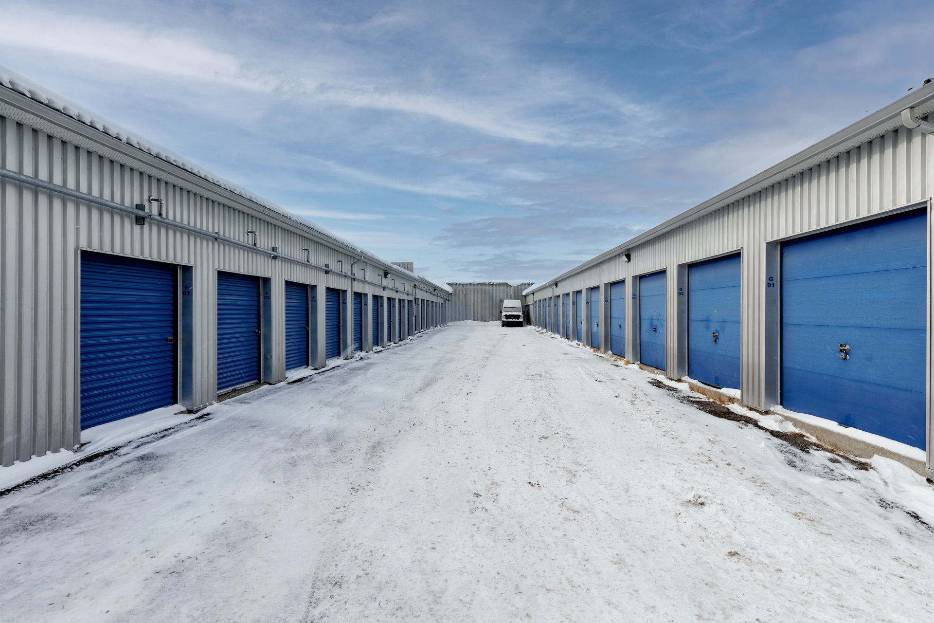 Des rangées de portes bleues de box de stockage, un chemin enneigé sous un ciel nuageux.