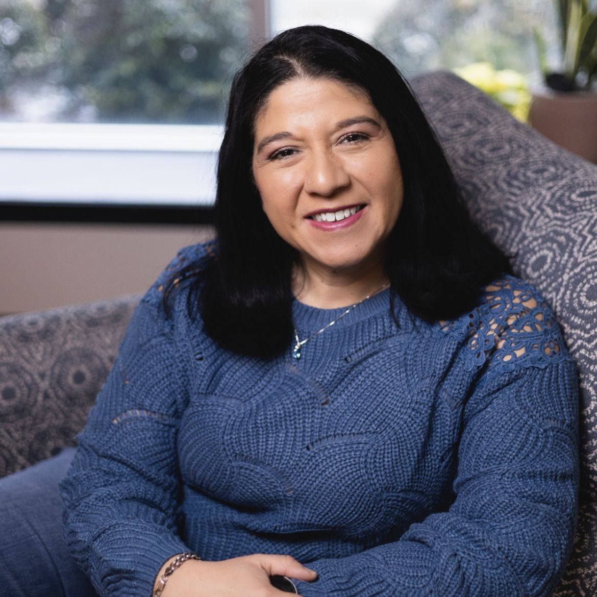 A woman in a blue sweater is sitting on a couch and smiling.