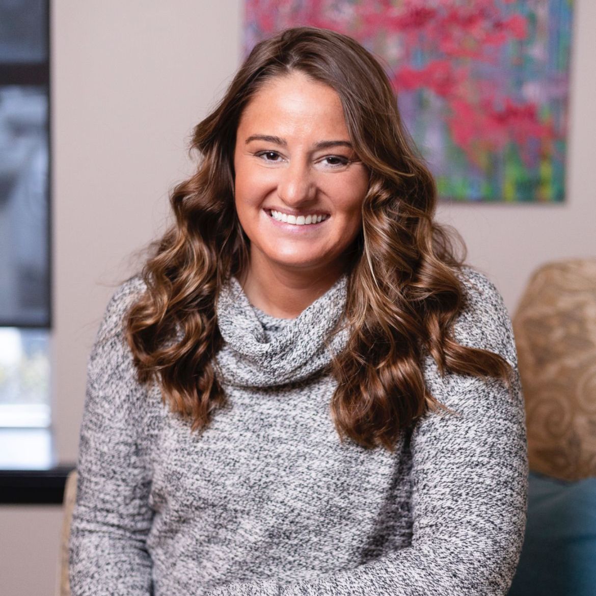 A woman in a grey sweater is smiling for the camera while sitting on a couch.