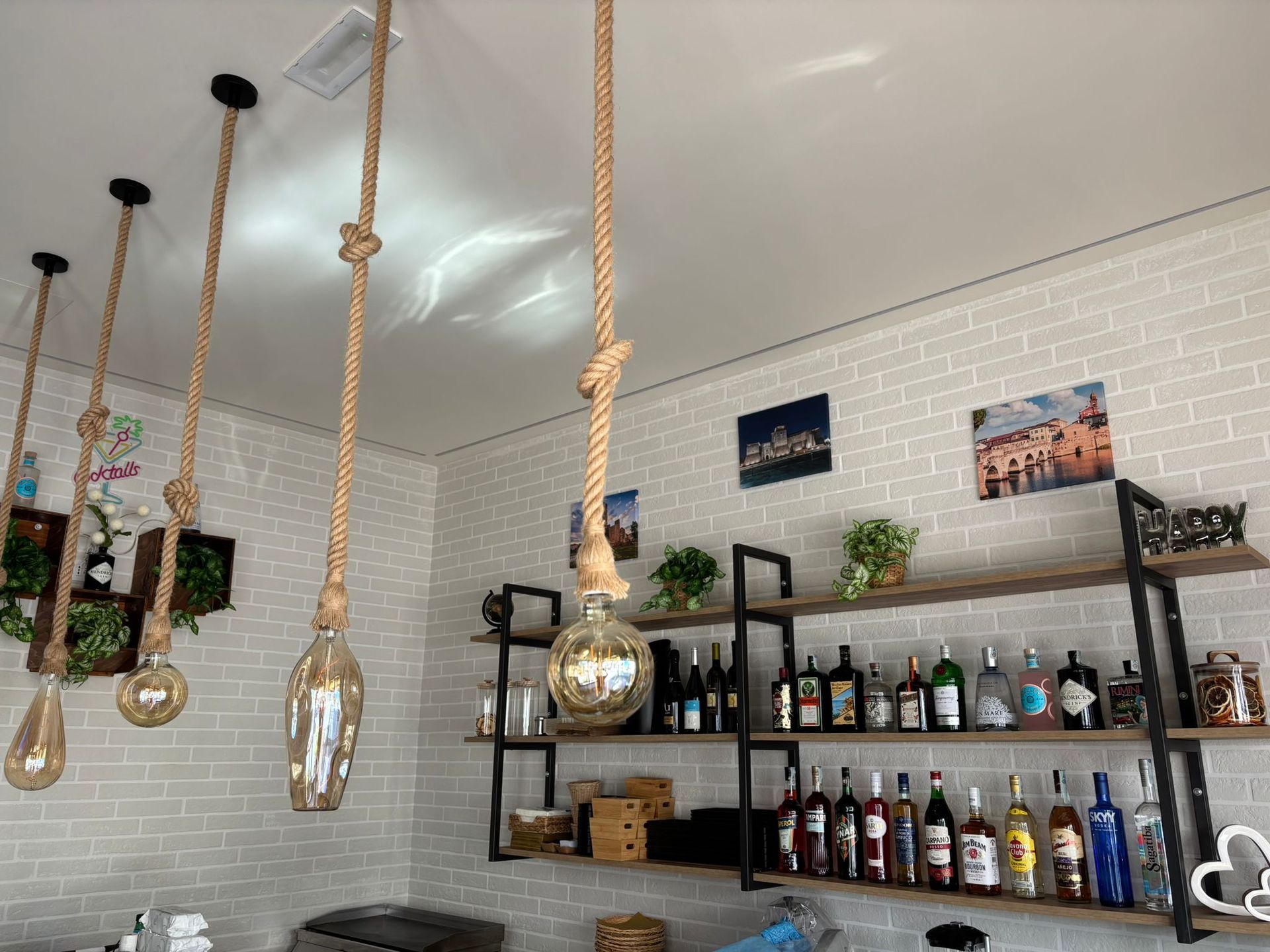 In un bar, delle rustiche lampade a sospensione in corda pendono sopra degli scaffali pieni di bottiglie di liquore, addossate a una parete di mattoni bianchi.