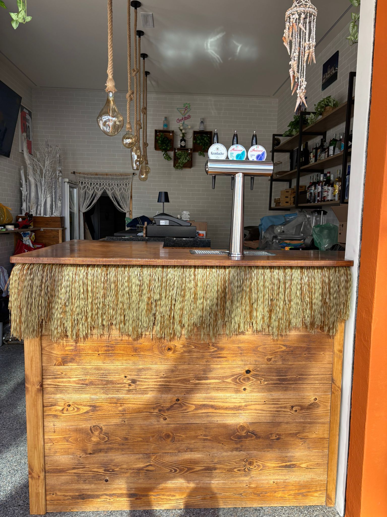 Un bancone da bar rustico in legno, ornato con frange di rafia color cuoio, dotato di spillatore di birra, lampade a sospensione e scaffali sul retro.