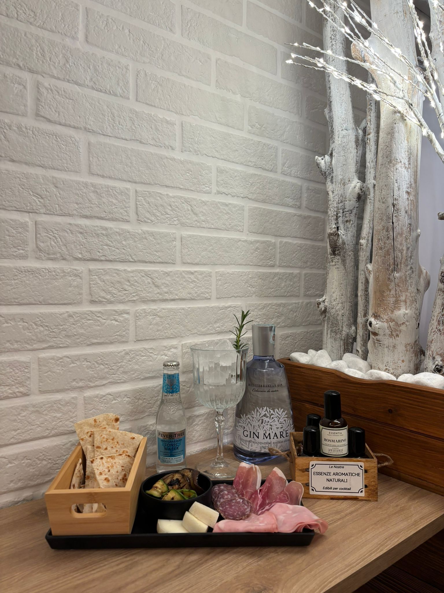 Un vassoio di antipasti con cracker, formaggi e salumi, accanto alle bevande, è disposto su un tavolo contro una parete decorativa in mattoni.