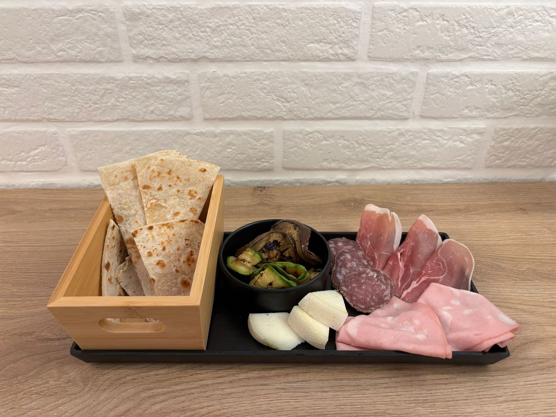 Un tagliere di salumi con affettati, formaggi, verdure grigliate e focaccia, presentato in una scatola di legno su una superficie di mattoni bianchi.