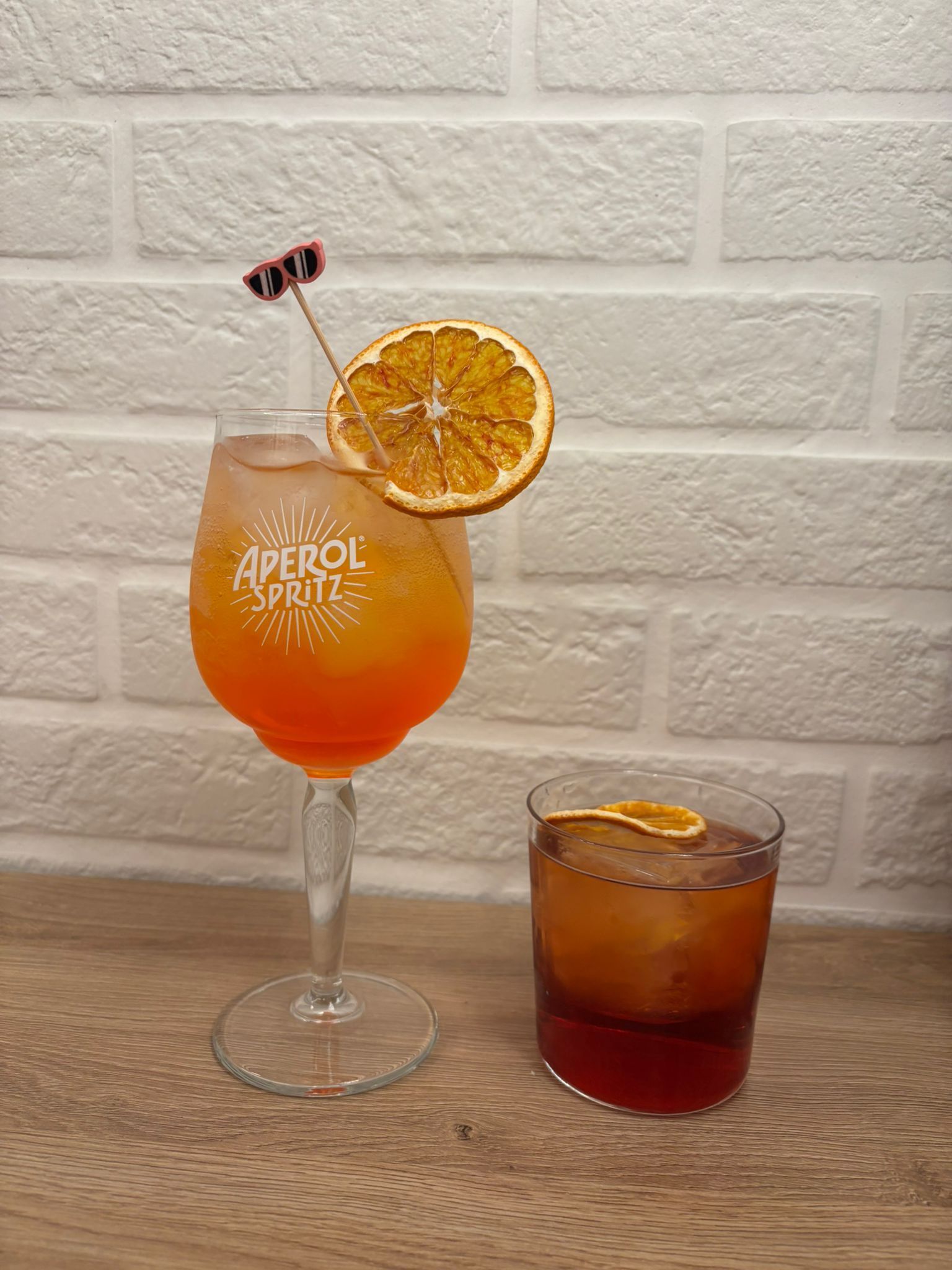 Un Aperol Spritz in un calice da vino di marca e un cocktail rosso scuro in un bicchiere old fashioned, entrambi guarniti con arancia essiccata.
