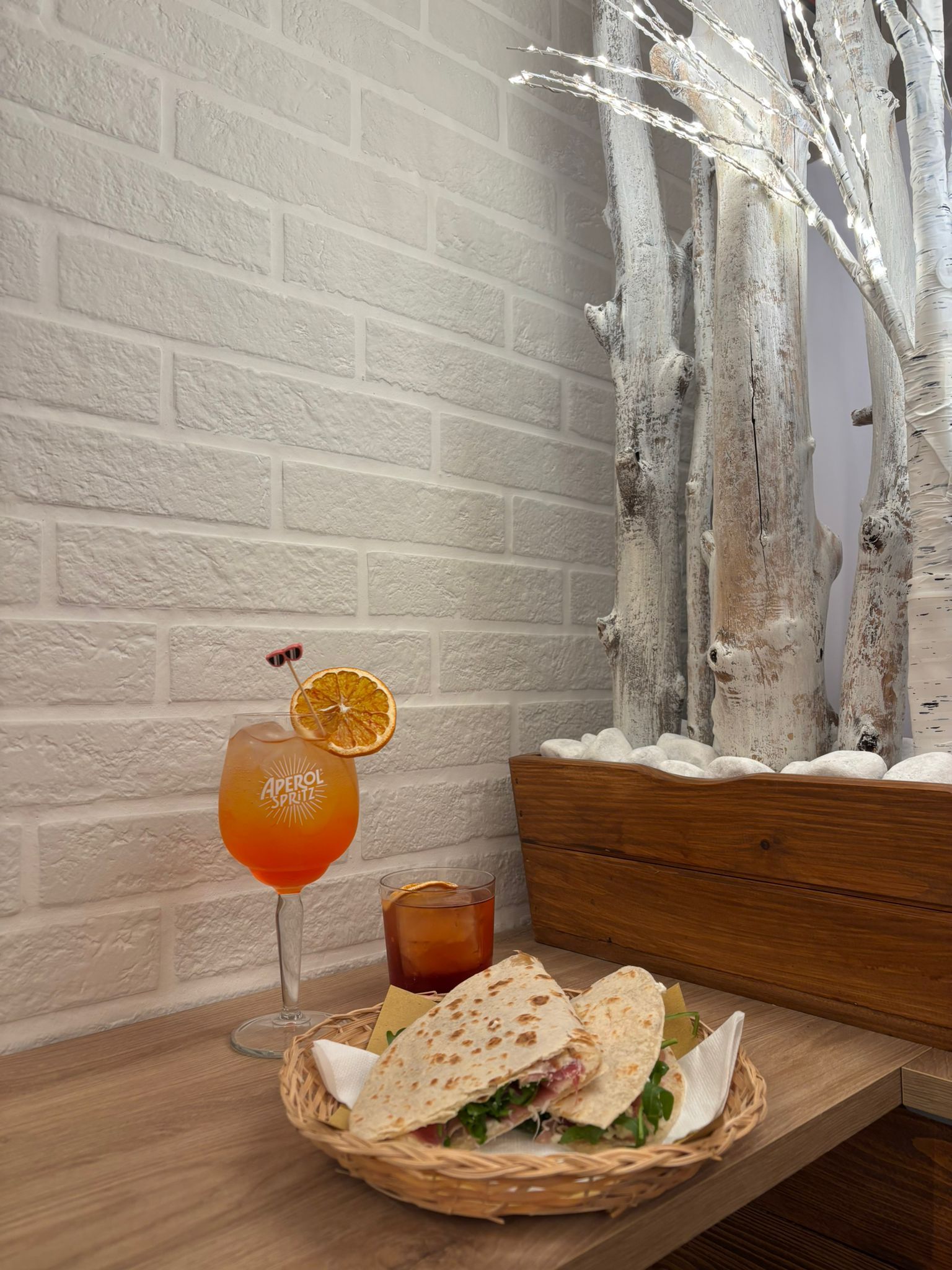 Un cocktail con una fetta d'arancia e un panino piegato in un cestino su un tavolo contro una parete di mattoni bianchi e tronchi decorativi.