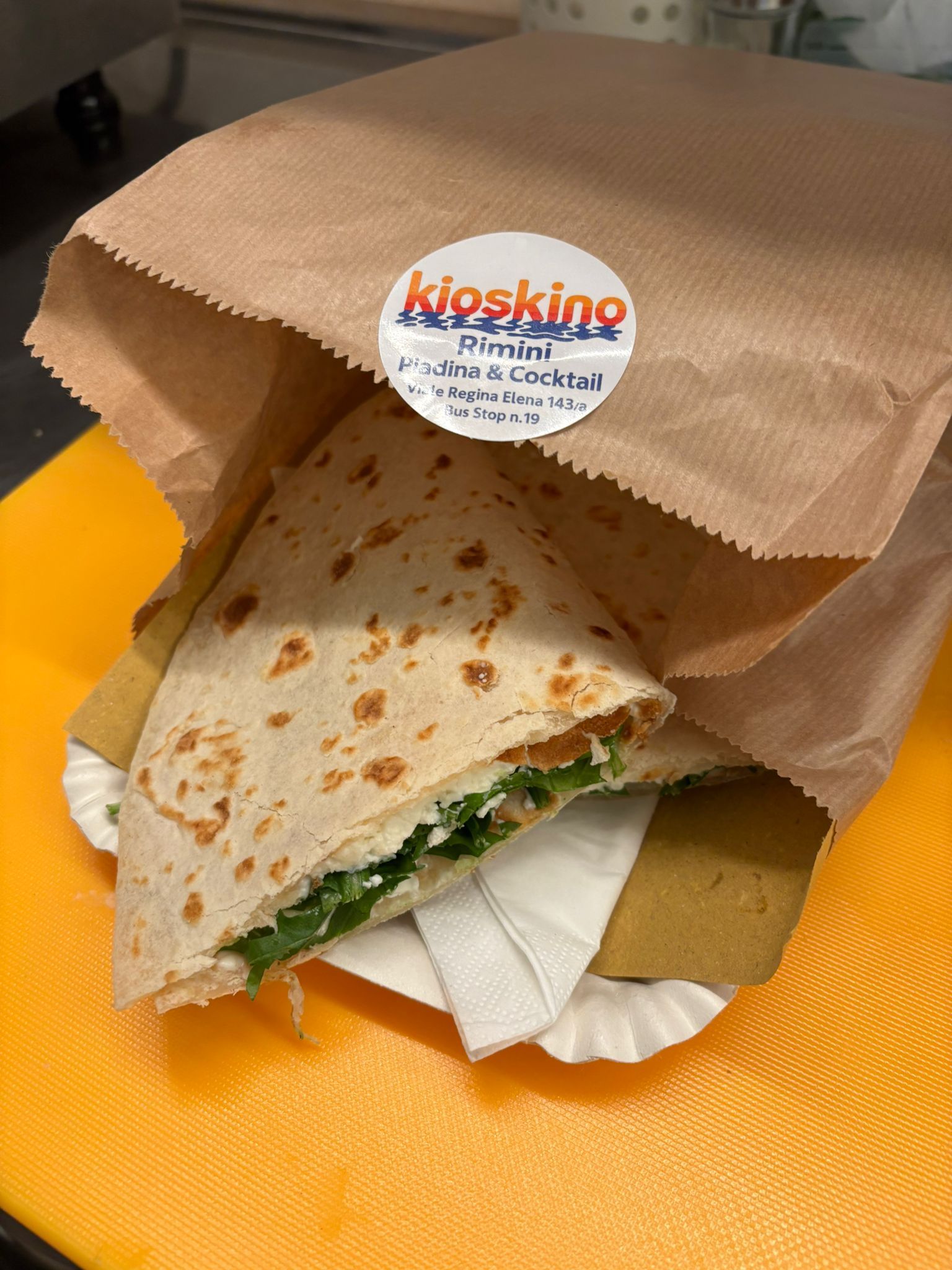 Un panino con pane piatto piegato, verdure e formaggio bianco, servito su un piatto di carta all'interno di un sacchetto di carta marrone.