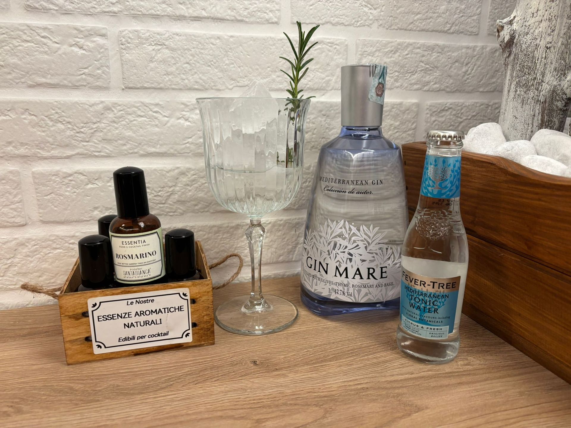 Un gin tonic preparato da Gin Mare su un piano di legno, completo di bottiglia di gin, acqua tonica, bicchiere pieno di ghiaccio e aromi.