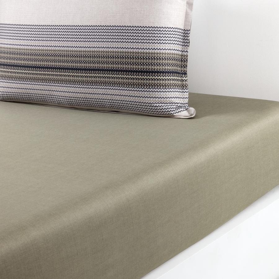 Un coin de matelas recouvert de draps vert olive, avec un oreiller à rayures grises et blanches posé contre un mur blanc.