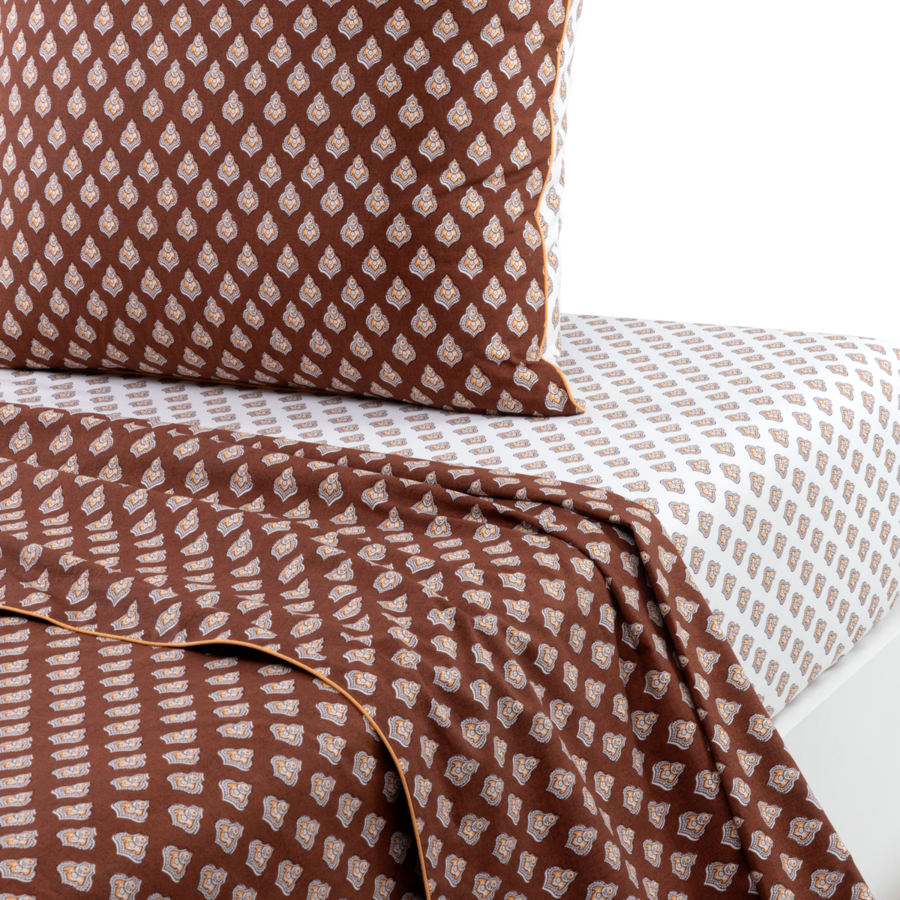 Un lit avec un oreiller marron et une couverture marron ornée d'un motif de feuilles blanc cassé, posés sur un drap blanc à motifs.