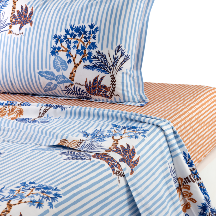 Gros plan sur une parure de lit à motifs botaniques rayés bleus et blancs, superposée à un drap à fines rayures orange.